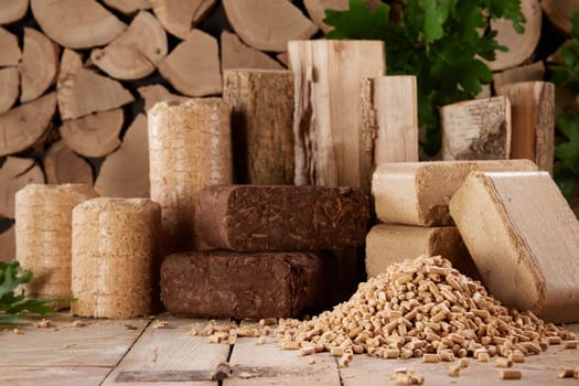 Various compressed biofuel briquettes and pyramid of wood pellets arranged on timber table against stack of firewood in daylight