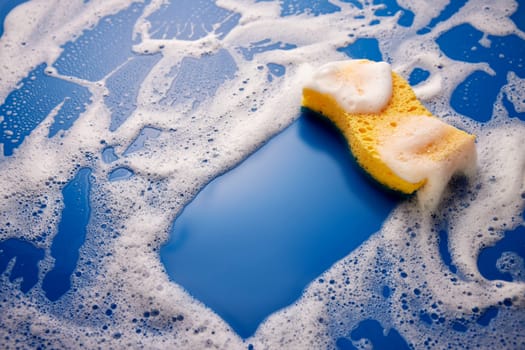 From above of yellow cleaning sponge placed on wet blue background covered with foam and soap bubbles during household routine