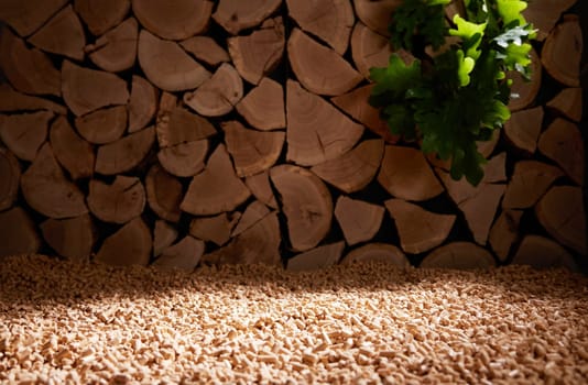 Green oak leaves hanging from pile of cut firewood stacked near scattered fuel pellets in daylight