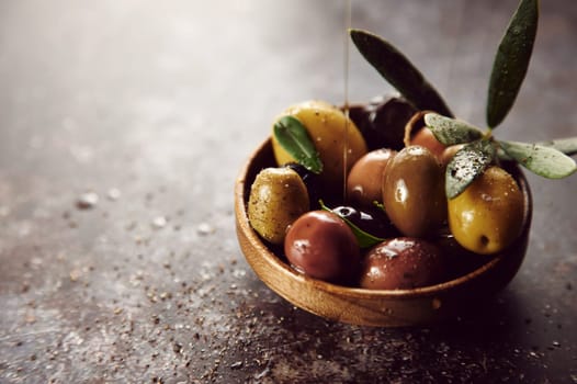 Olive oil jet pouring on mix of tasty marinated olives in bowl placed on gray background with seasoning in light kitchen
