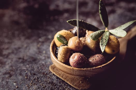 Jet of olive oil pouring on appetizing assorted marinated olives in wooden bowl placed on gray background in light kitchen