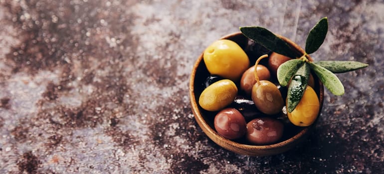 From above mix of marinated delicious olives in bowl with leaves placed on gray table in light kitchen