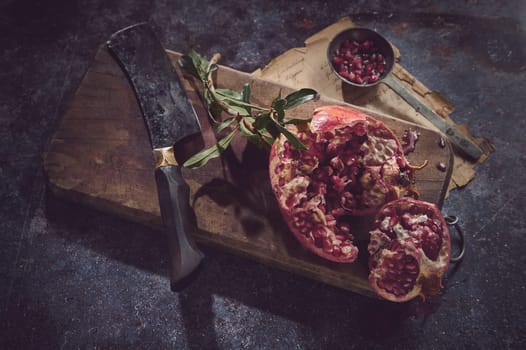 From above of fresh red pomegranate served on wooden chopping board with cleaver knife on gray background with old paper
