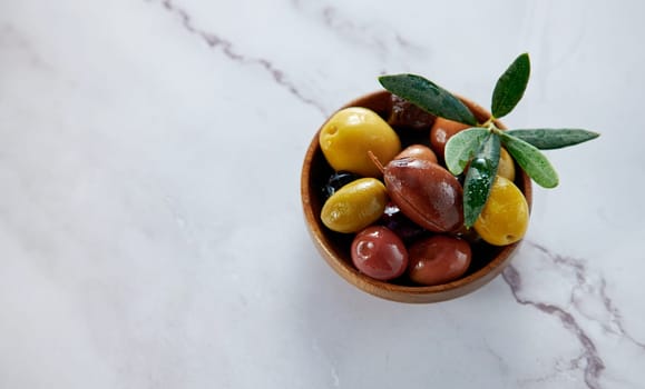 From above of tasty fresh different types of ripe marinated olives with green leaves served in bowl on white background