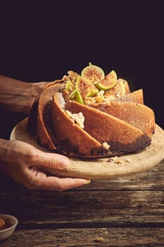 Crop anonymous cook holding sweet homemade freshly baked fig cake served on wooden cutting board above table in light kitchen
