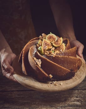 Crop unrecognizable cook holding sweet freshly baked cake made of figs with decorations on wooden cutting board in light kitchen