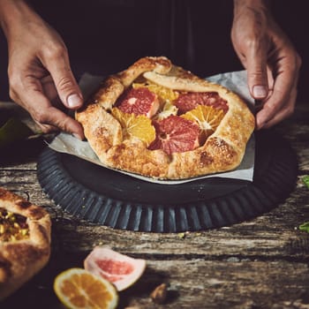 Hands of crop anonymous cook holding tasty sweet homemade freshly baked tart with citrus fruits at wooden table in light kitchen