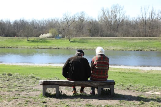 Two seniors enjoy the warming spring sun on a wooden bench on the banks of the Rhine near Hartheim.