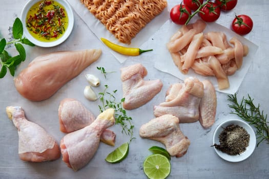 Top view of different types of raw chicken meat placed on gray background with greens and condiments in light kitchen