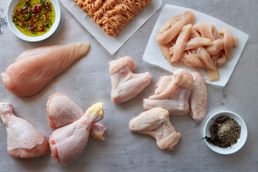 Top view of raw chicken mince and breasts placed near drumstick and wings on gray background with uncooked strips and flavoring