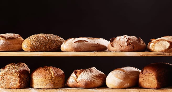 Abundance of freshly baled loaves of delicious bread with crispy crusts placed on wooden shelves against black background in light studio