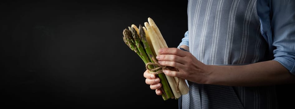 Crop anonymous female cook with uncooked healthy green and white asparagus standing on black background in studio during cooking preparation
