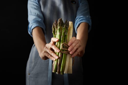 Crop anonymous female holding fresh heap of fresh uncooked raw and white asparagus while standing against black background in studio