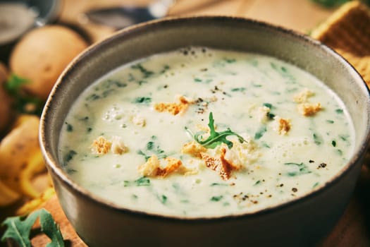 Tasty fresh cream roucola soup with croutons served on table in light kitchen with potatoes against blurred background at home