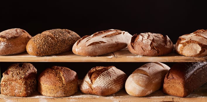 Bunch of appetizing freshly baked loaves of bread placed on wooden shelves with flour against black background in light studio