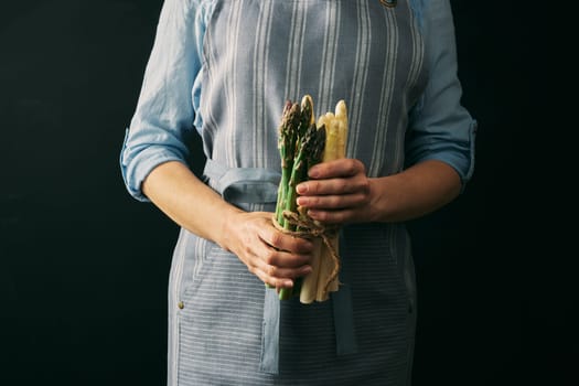 Crop faceless female cook in apron holding mix of raw healthy asparagus against black background during cooking preparation in studio