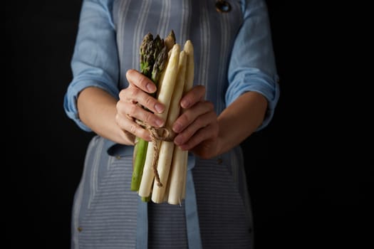 Crop unrecognizable female cook in apron holding bunch of raw healthy white and green asparagus against black background in studio