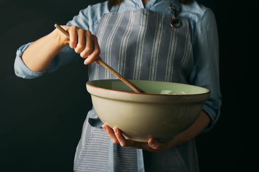 Crop anonymous female cook in apron mixing ingredients into bowl with wooden spatula while preparing for baking against black background