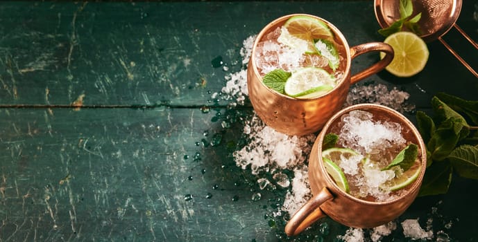 High angle of tasty alcoholic Moscow mule drinks with ice and lime served on dark wooden table with mint in light kitchen