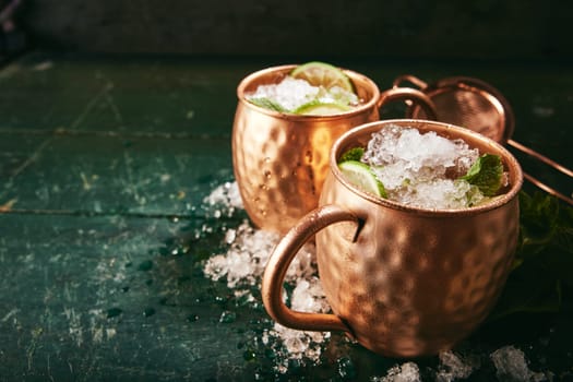 Refreshing alcoholic Moscow mule drinks with lime slices in copper cups placed on shabby table with ice in light kitchen