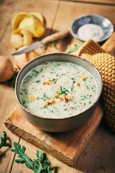 From above of delicious cream rucola soup served on wooden table with peeled potatoes and sour cream in light kitchen