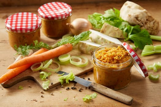 Glass jars filled with fresh vegetable conservation placed on wooden table with carrots and celery in light kitchen during cooking process