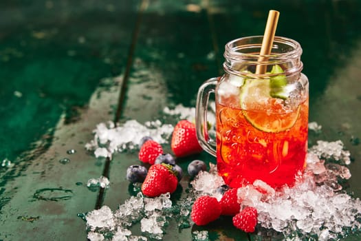 Glass jar filled with red berry beverage with lime served with straw on table with ice and strawberries and blueberries