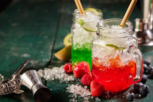 Refreshing tasty red and green beverages served with ice and straws in glass jars on table with assorted berries and jigger