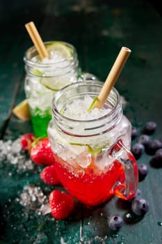 From above of glass jars filled with red and green berry beverages served with straws on table with strawberries and blueberries in kitchen