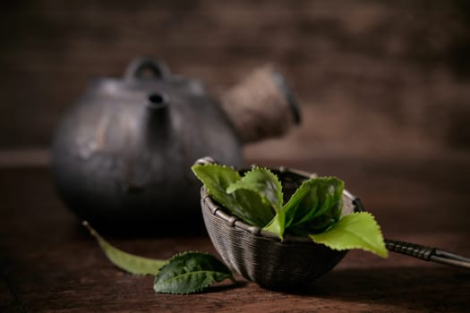 Fresh green tea leaves in ladle placed on table near old fashioned metal teapot on blurred background in light room