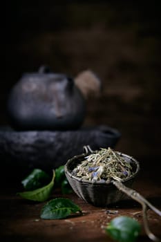 Aromatic loose tea and fresh green leaves placed on table with old fashioned metal teapot on blurred background in room