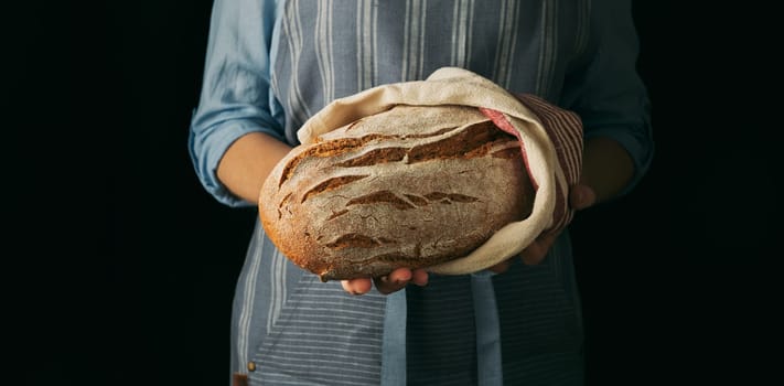 Crop anonymous female baker in apron holding freshly baked loaf of sourdough bread wrapped in towel against black background in modern studio