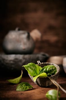 Green aromatic leaves of tea in ladle placed on wooden table with vintage metal teapot against blurred background in room