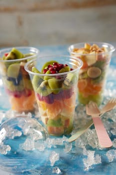 Mix of fresh tasty chopped fruits in plastic cups place don table with ice and wooden forks in light room