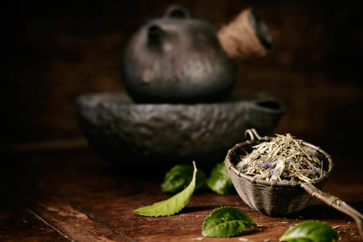 Heap of loose tea in ladle on wooden table with aromatic green leaves near old metal teapot in light kitchen