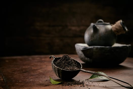 Pile of aromatic loose tea for brewing placed on wooden table with fresh green leaves and old teapot in kitchen