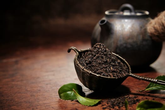 Heap of dried loose tea in metal ladle placed on wooden table with tea teapot in kitchen during teatime preparation