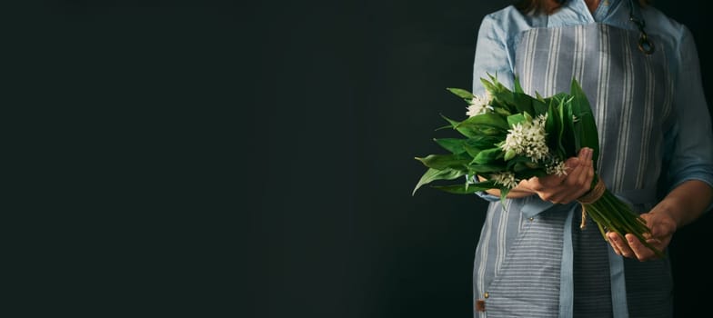Crop anonymous female cook in apron holding wild garlic with green leaves while standing against black background during cooking preparation