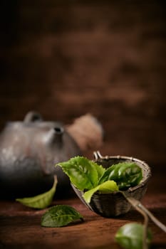 Green tea leaves for brewing in metal ladle placed on table with old teapot on blurred background in light room