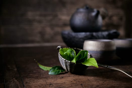 Fresh green aromatic tea leaves in ladle placed on wooden table with old fashioned teapot and cups in light room