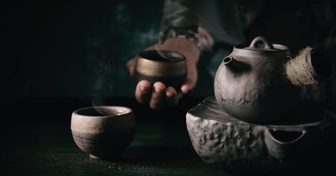 Crop unrecognizable person holding cup of steaming hot tea near table with old fashioned metal teapot in kitchen during teatime