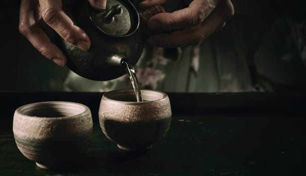 Crop faceless person pouring hot drink from old fashioned metal teapot into cup on table during tea ceremony in kitchen