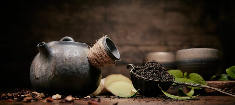 Dried loose tea near metal vintage teapot and cups placed on table with various scattered condiments and green leaves in kitchen