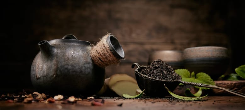 Aromatic loose tea near old fashioned teapot placed on table with scattered condiments and ginger in kitchen with set of cups