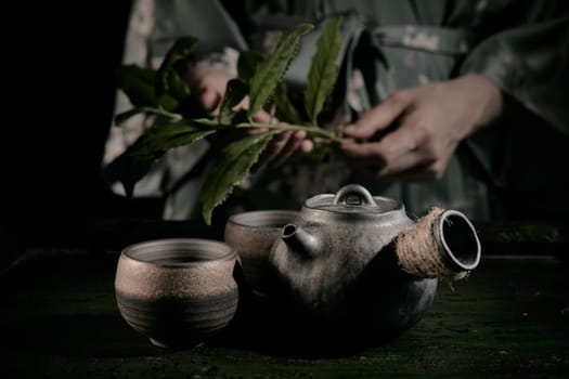 Crop unrecognizable person with fresh green tea leaves standing at table with retro metal teapot and cups in kitchen