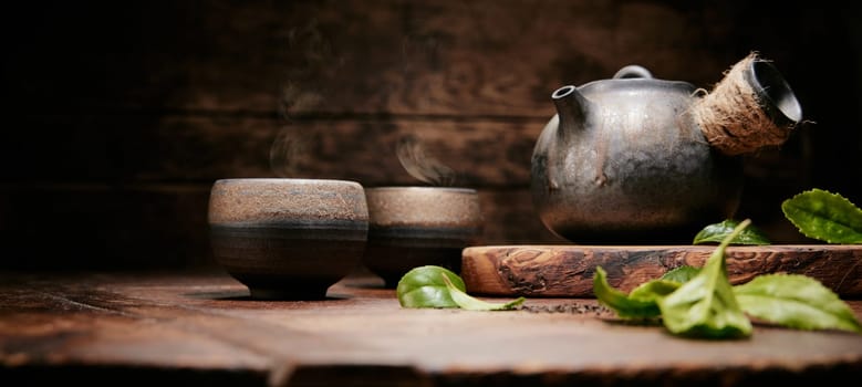 Green fresh tea leaves for brewing on wooden table with tea cup and retro metal teapot in kitchen during tea time