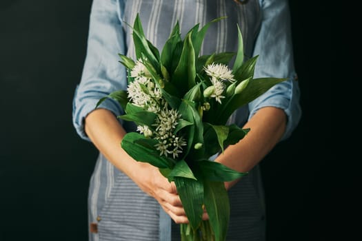 Crop anonymous female cook in apron holding heap of wild garlic with white bulbs and green leaves against black background