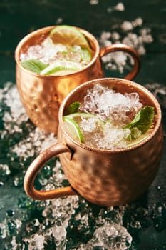 From above of alcoholic Moscow mule beverages with lime slices served in copper mugs on table with ice in kitchen