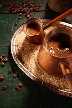 From above of metal cezves with freshly brewed Turkish coffee on tray with sand placed on table with scattered coffee beans