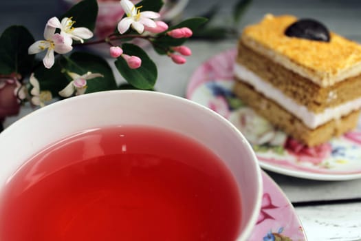 A cup of tea large smooth and a cake on a wooden table with flowers in the background..